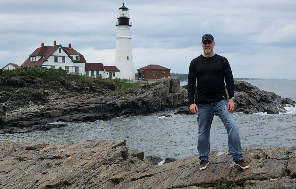 A man stands on cliffs in front of a lighthouse on the ocean.